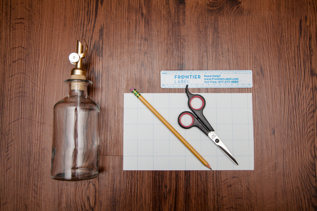 Glass bottle, blank graph paper, pencil, scissors, and ruler assembled on a wooden table