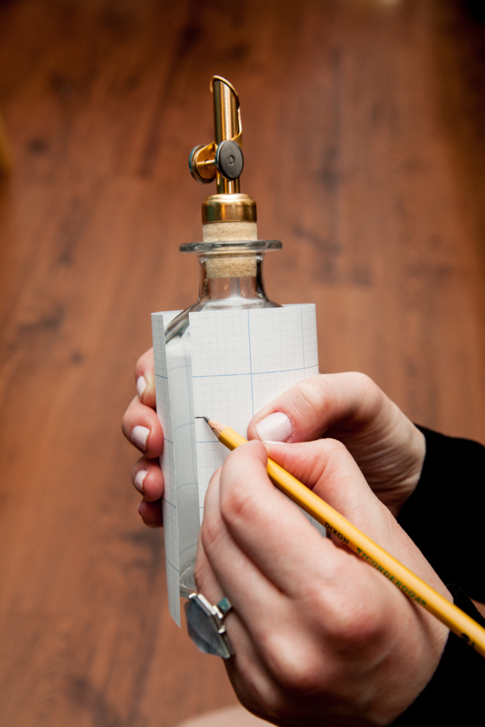 Woman holding graph paper around a glass jar and marking label edges.
